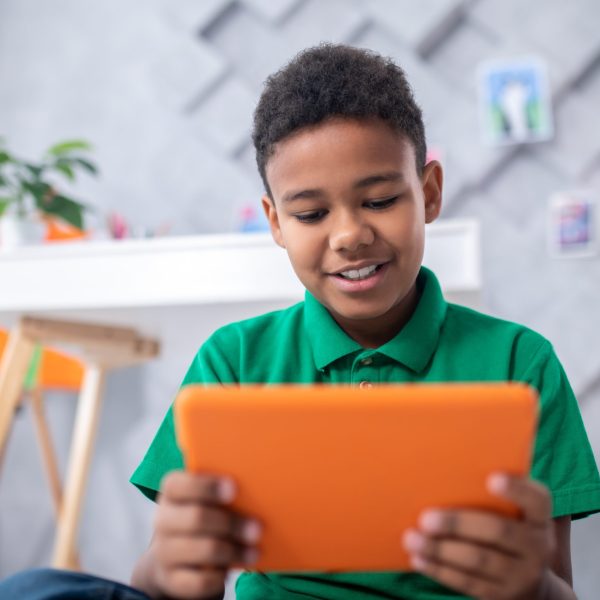 Interesting information. Dark-skinned boy of primary school age in green tshirt looking with interest at orange tablet sitting in room with table and paintings